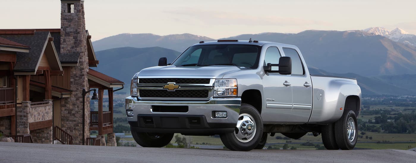 A silver 2014 used Chevy Silverado 3500 is parked next to a stone cabin with mountains in the distance.