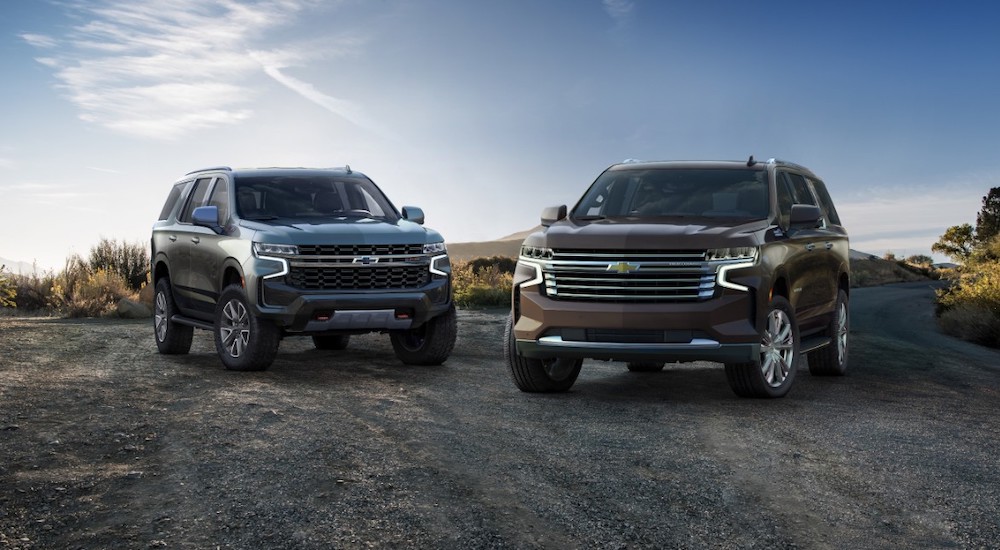 A grey 2021 Chevy Tahoe is next to a brown 2021 Suburban in a rural parking area in front of a blue sky.