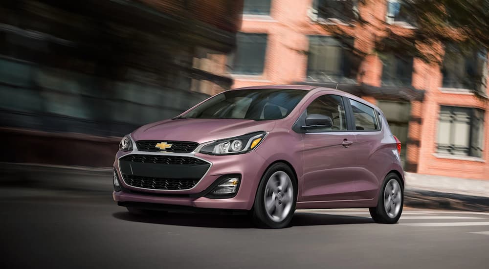 A pink 2021 Chevy Spark is driving past a brick building after leaving a Chevy dealer.