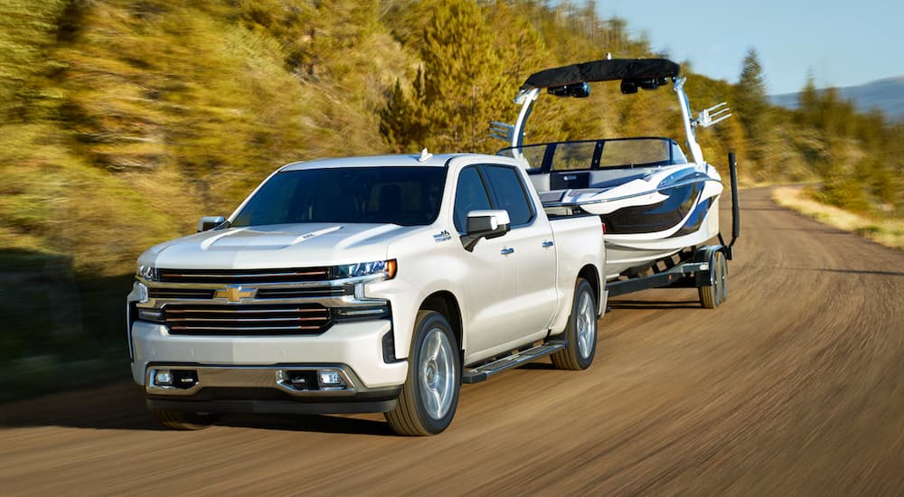 A white 2021 Chevy Silverado 1500, popular among Chevy trucks, is towing a boat past trees.