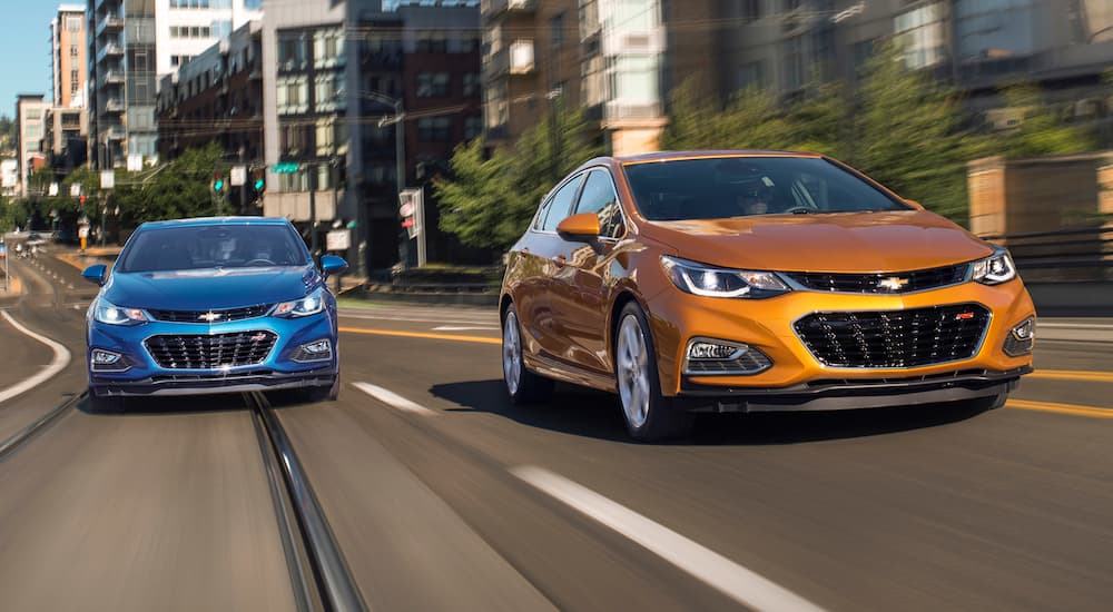 An orange and a blue 2017 Chevy Cruze are driving on a city street with multiple lanes.