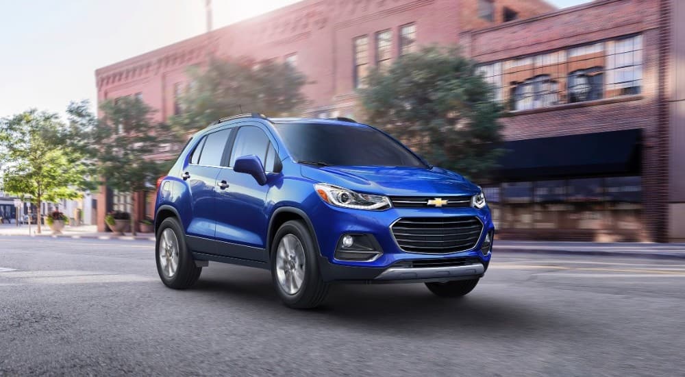 A blue 2017 Chevy Trax is driving on a city street past a brick building.