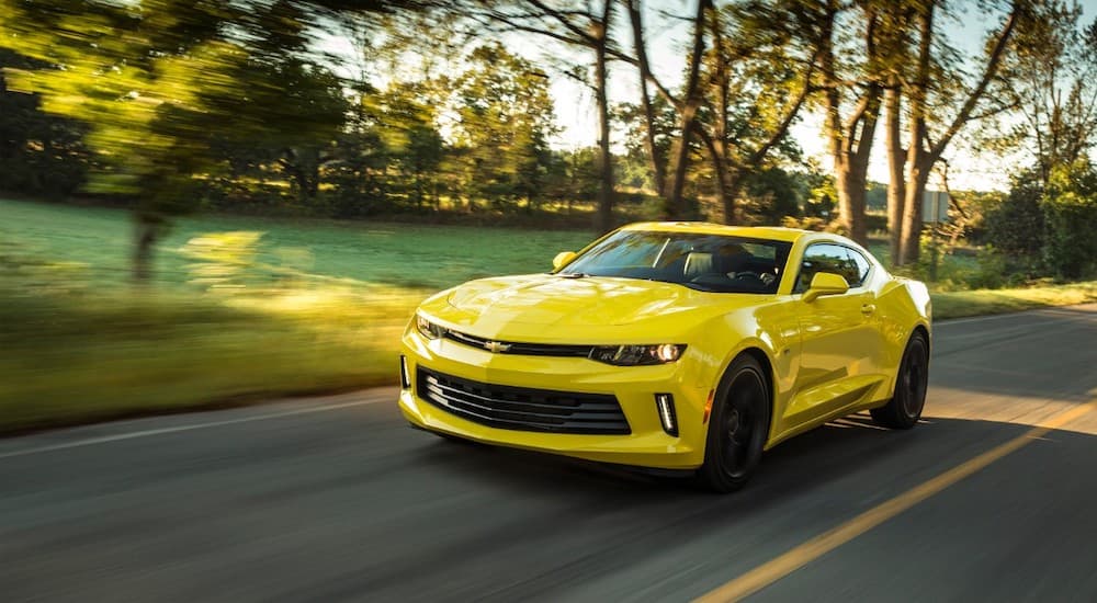 A yellow 2018 Chevy Camaro is driving on a sunny street past trees after leaving a used car dealer.