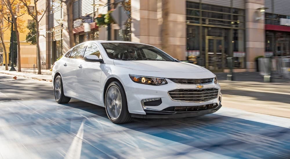 A white 2018 Chevy Malibu is driving through a city intersection after leaving a used car dealer.