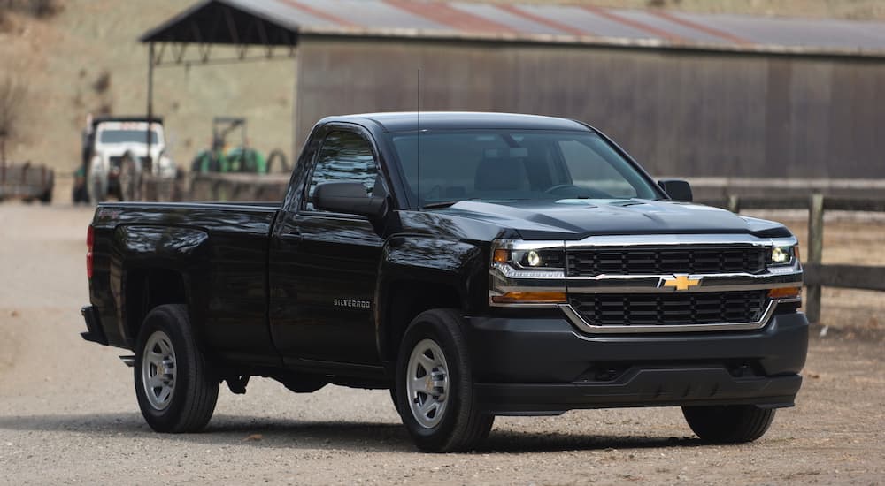 A black single cab 2016 used Chevy Silverado is parked in front of a dump truck and metal storage building.