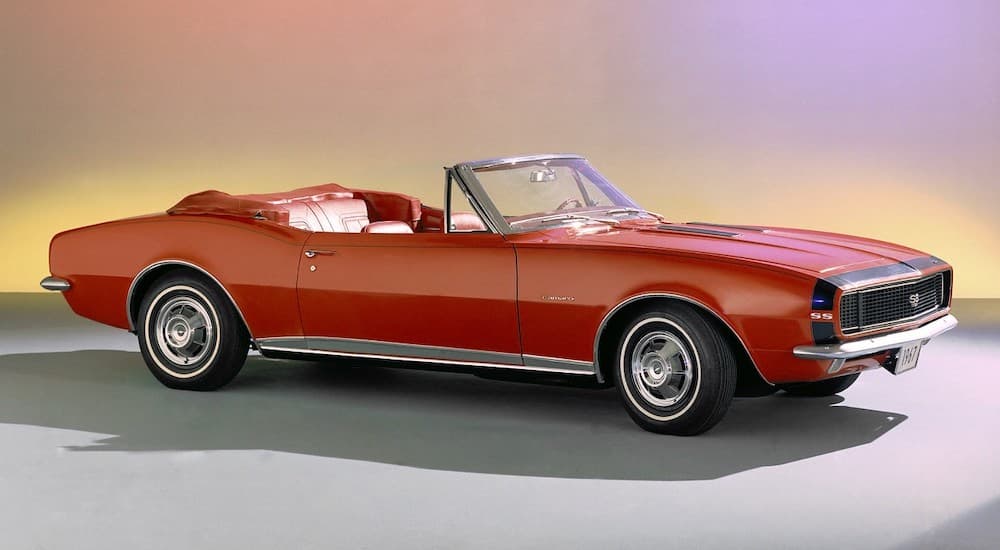 A red 1967 Chevy Camaro convertible is parked in a showroom with a multi-color background.