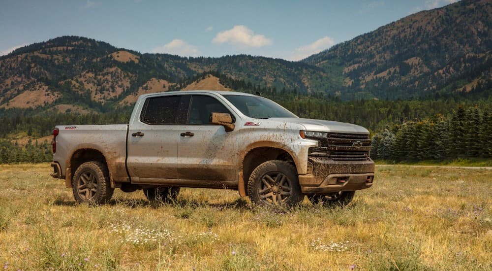A silver 2019 Chevy Silverado 1500 is covered in mud in a field in front of hills.