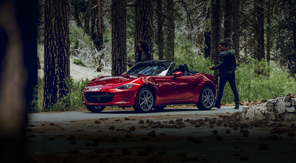 A couple is shown approaching a red 2020 Mazda MX-5 Miata parked in the woods.
