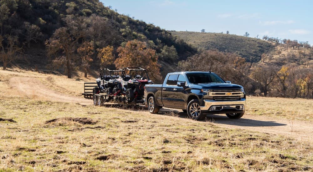 A black 2020 Chevy Silverado 1500 is shown parked near a trail towing two UTVs.