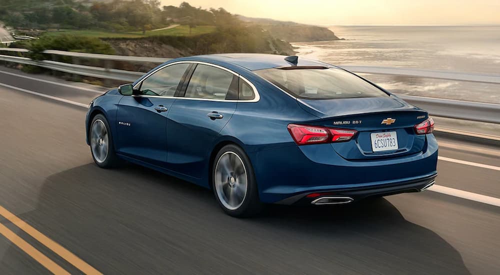 A blue 2022 Chevy Malibu is shown from a rear angle driving past an ocean.