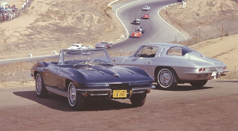 A blue and a silver 1966 Chevy Corvette Stingray are shown parked next to a racetrack.