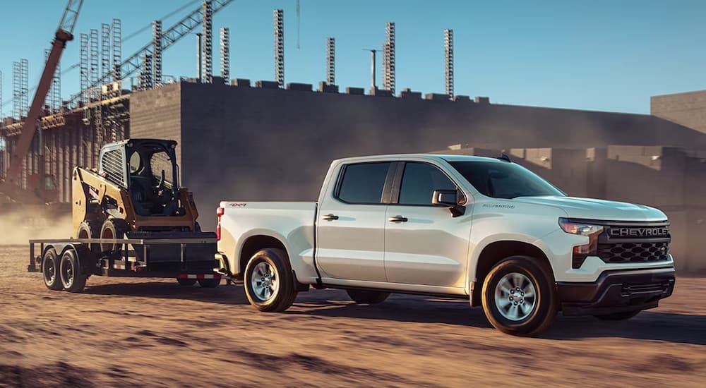 A white 2023 Chevy Silverado 1500 Custom is shown from the side while towing a skid-steer.