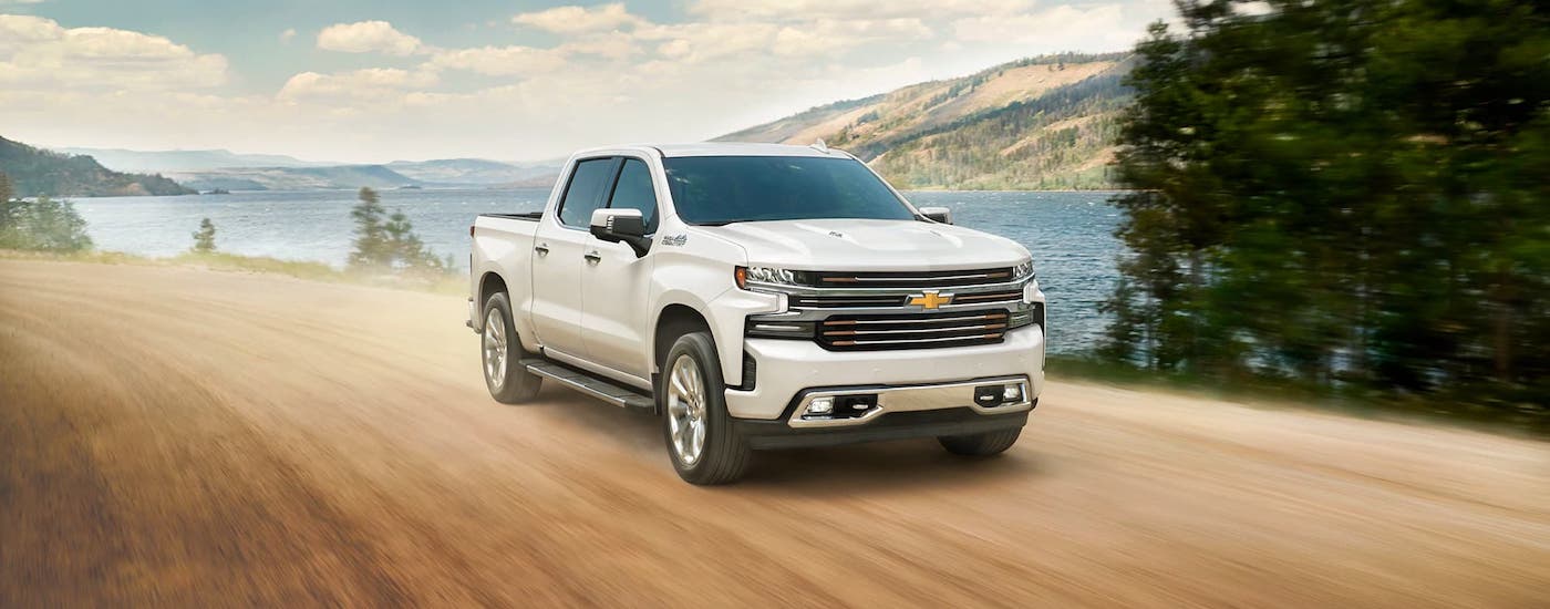 A white 2021 Chevy Silverado 1500 is driving on a dirt road past a lake and distant mountains.