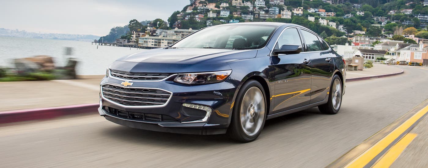 A blue 2019 Chevy Malibu is driving past a body of water and away from a distant hill covered with houses.