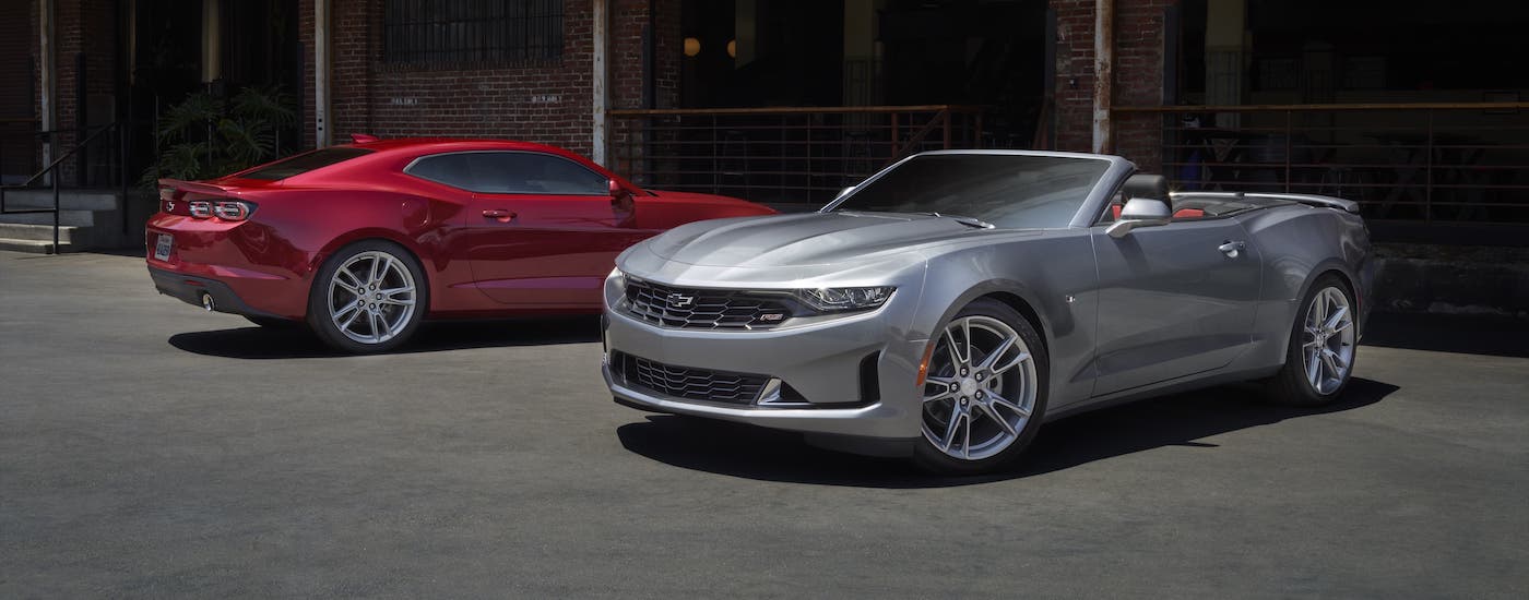 A red coupe and a silver convertible 2021 Chevy Camaro from your local Chevy dealer are parked in front of a brick building.