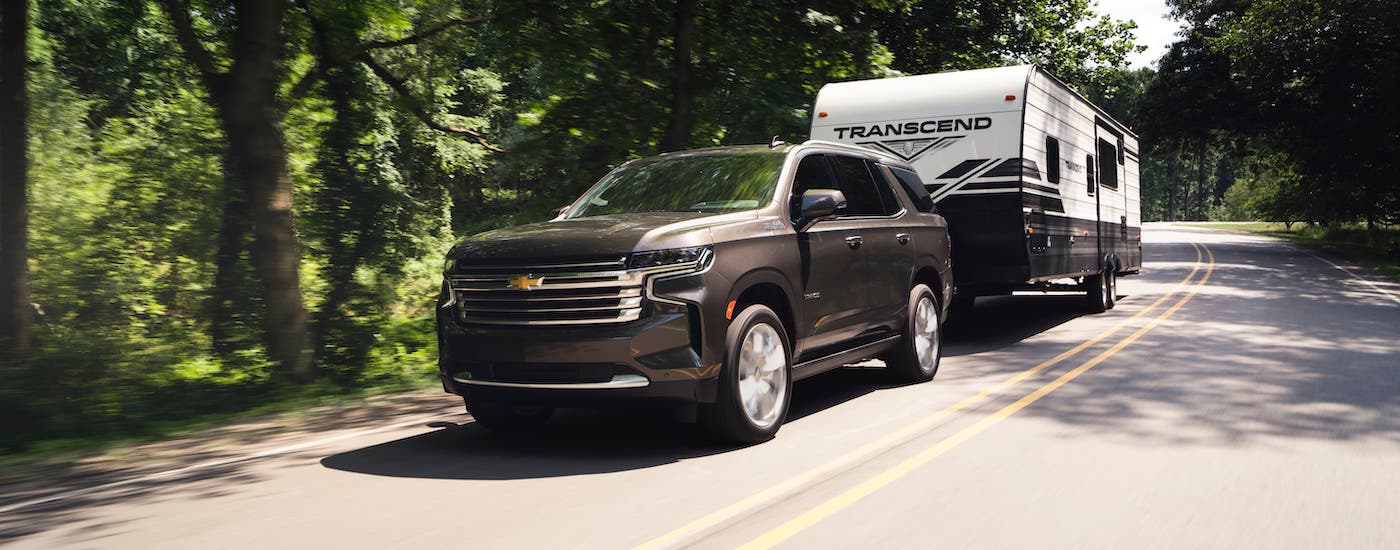 A brown 2021 Chevy Tahoe High Country is towing a camper on a tree-lined road.