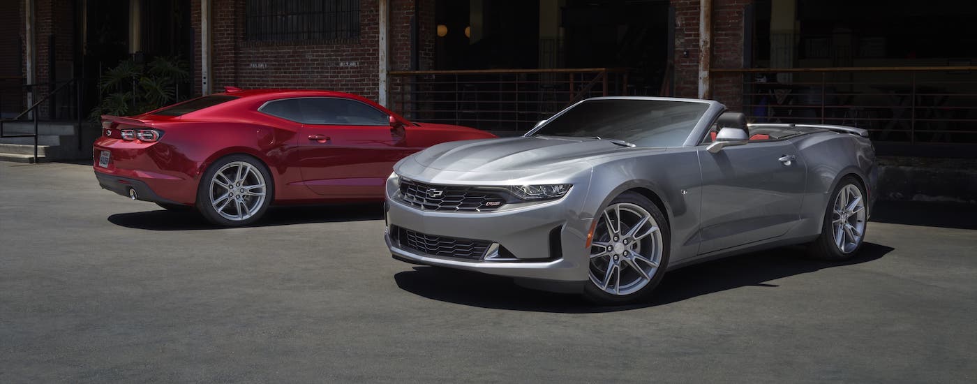 A silver 2021 Chevy Camaro convertible and a red coupe are parked in front of a red brick building outside a Chevy dealer in Houston.