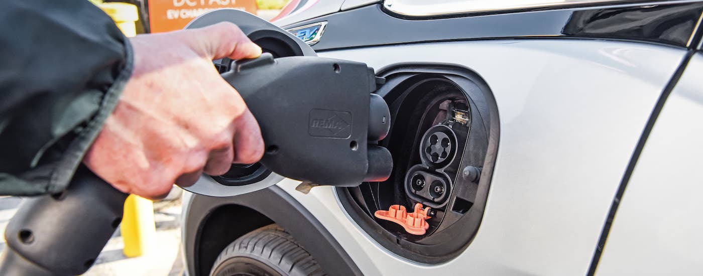 A closeup shows a hand plugging in the charging cord of a popular Chevy EV, a silver 2021 Chevy Bolt EV.