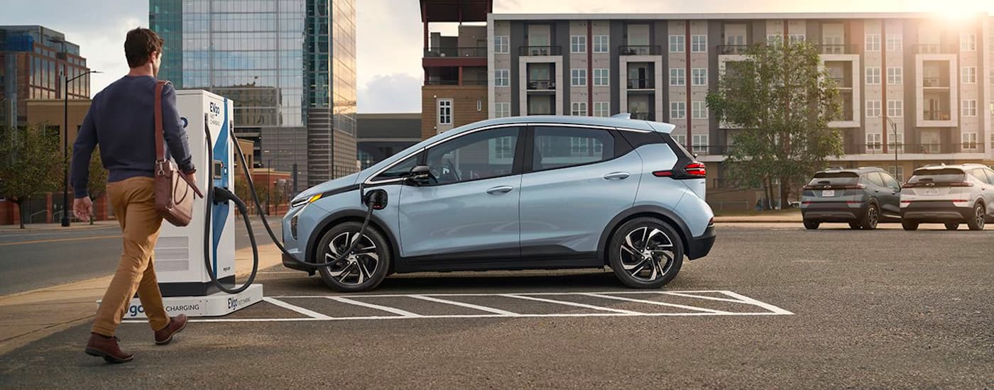 A man is shown approaching a light blue 2022 Chevy Bolt EV at a charging station after leaving a Pinehurst Chevy dealer.
