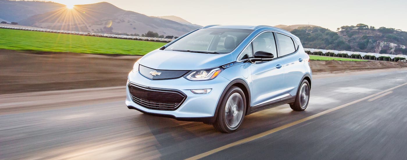 A silver used Chevy EV, a 2018 Bolt EV, is driving in front of a field.