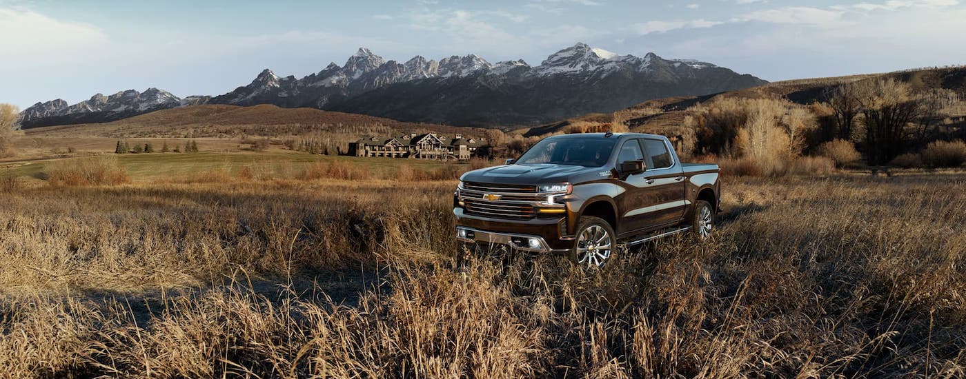 A brown 2019 Chevy Silverado High Country is parked in a field in front of a mansion and snow-covered mountains.