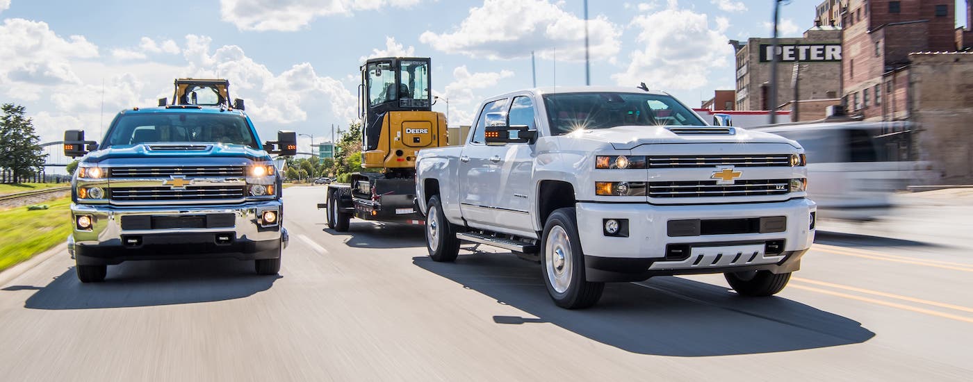 A blue and a white 2018 Chevy Silverado 2500 are driving on a highway towing construction equipment and shown from the front.