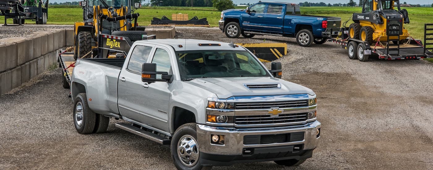 A silver 2018 used Chevy Silverado 3500 and a blue Silverado 2500 are parked on gravel and towing trailers with construction equipment.