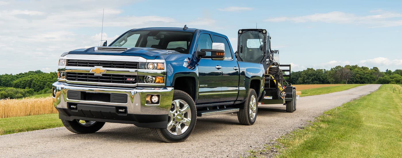 A popular used Chevy truck in Houston, a blue 2018 Chevy Silverado 2500HD, is towing construction equipment on a dirt road.