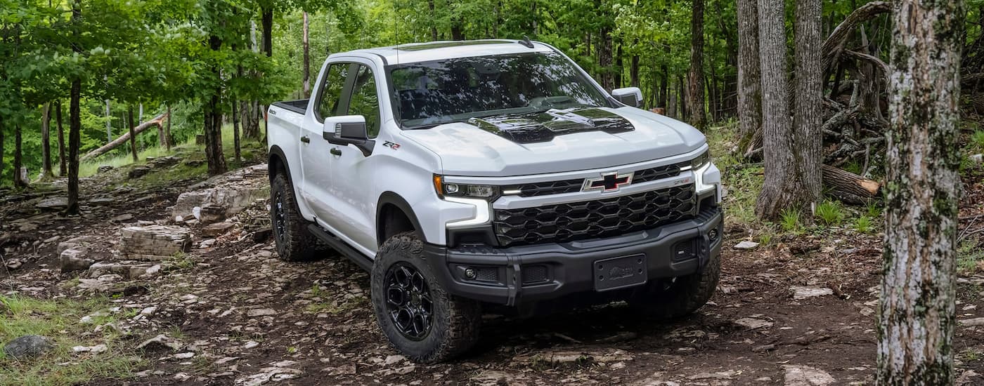 A white 2023 Chevy Silverado 1500 ZR2 Bison is shown driving on a rocky trail in a wooded area.
