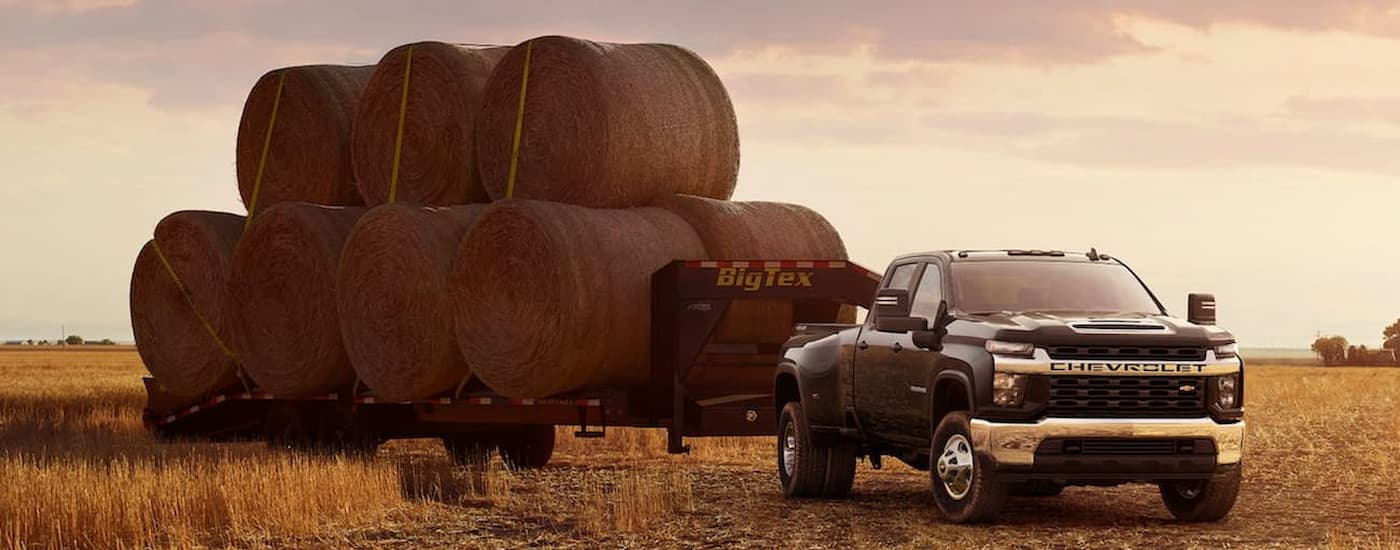 A black 2021 Chevy Silverado 3500HD is shown towing a large trailer of hay bales in an open field.